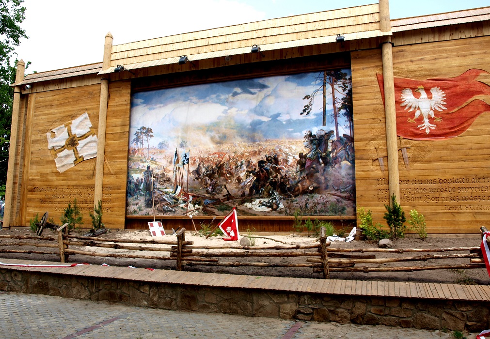Obraz przedstawiajacy bitwe wojsk pod Grunwaldem. osadonyy w specjalnej teatralnej konstrukcji z drewna. Po lewo i prawo prztwierdzone dwie flagi.