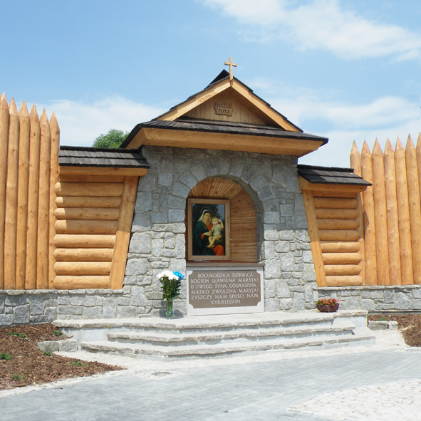Kaplica z białych kamieni. Przykryta drewnianym dachem. Z boku drewniana obudowa. W środku obraz.