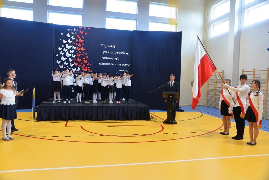 zdjęcie przedstawia wnętrze sali gimnastycznej w PSP Cerekiew. Na scenie stoją dzieci. Po prawej stronie znajduje się poczet sztandarowy z flagą