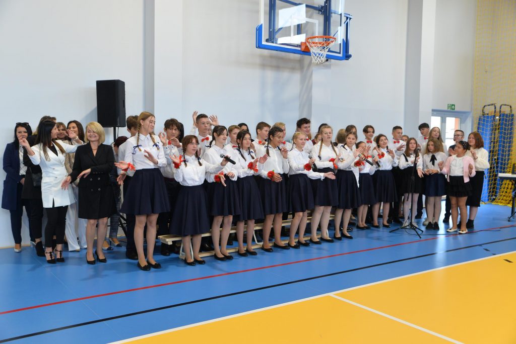 zdjęcie przedstawia wnętrze sali gimnastycznej w PSP Cerekiew. Widać młodzież wraz z nauczycielami śpiewającą piosenkę, wszyscy są uśmiechnięci i mają uniesione ręce. 