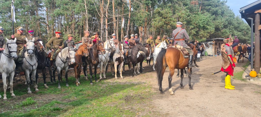 Zdjęcie na pierwszym planie przedstawia jeźdźców na koniach, którzy są ubrani 
w stroje kawaleryjskie i pochodzą z różnych stron Polski – trwają przygotowania 
do pokazów kawaleryjskich. W tle widać las.
