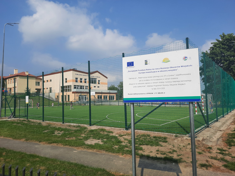 na zdjęciu boisko szkolne a przed min tablica informacyjna o dofinansowanie