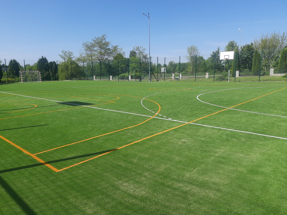 na zdjęciu boisko szkolne w zbliżeniu: na sztucznej nawierzchni namalowano pasy a w tle widać bramkę do gry w piłkę nożną oraz kosz do gry w koszykówkę
