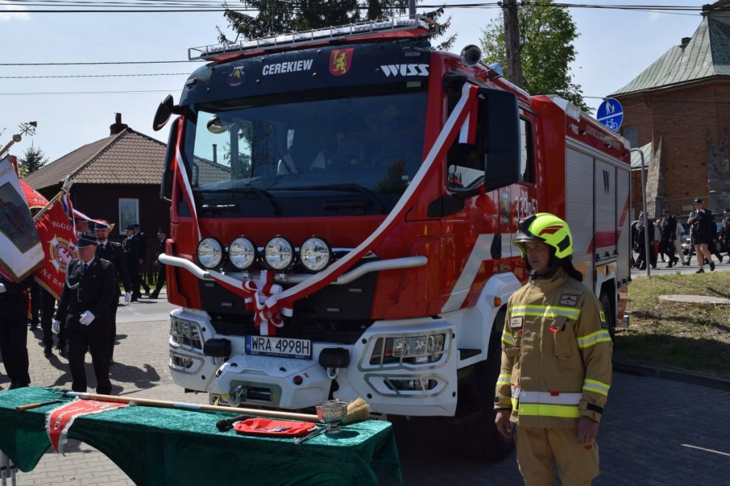 Uroczyste poświęcenie wozu strażackiego w OSP Cerekiew. W centralnym punkcie widać wóz strażacki przepasany biało-czerwoną wstążką. Po bokach stoją przedstawiciele straży pożarnej.