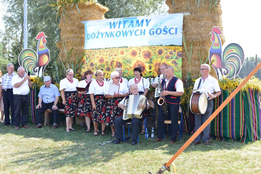 Kapela ludowa - wszyscy w strojach ludowych, niektórzy trzymają instrumenty muzyczne. w tle dekoracją ze słomy i baner Witamy dożynkowych gości.