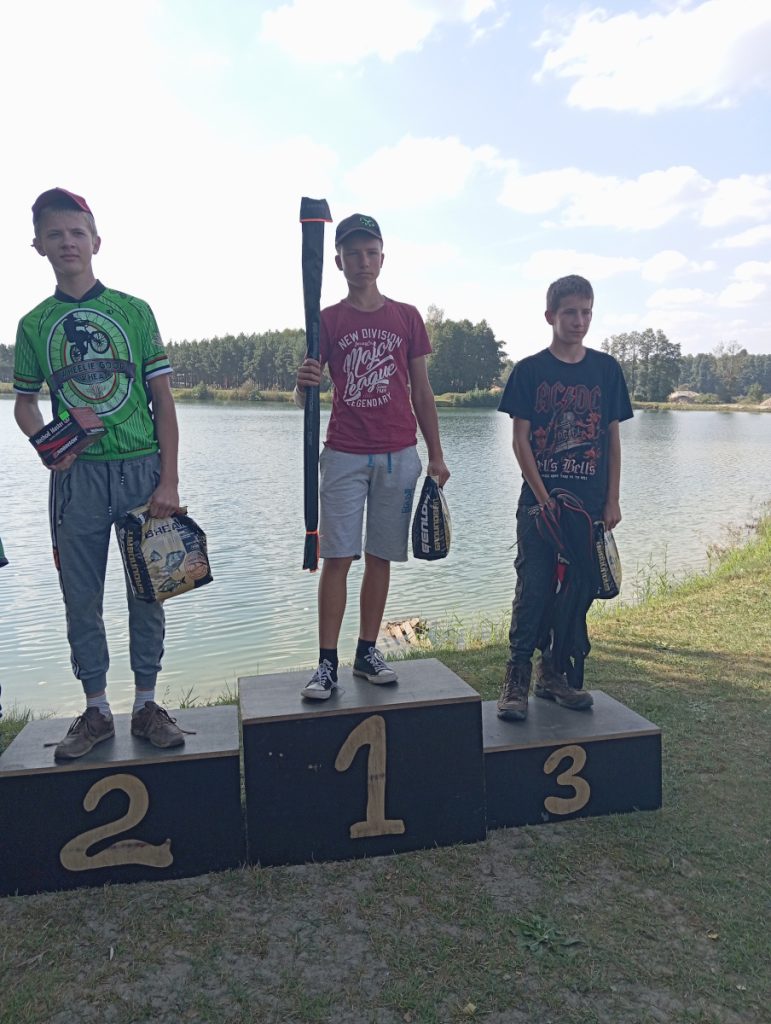 Na podium zwycięzcy zawodów wędkarskich wśród juniorów. Trzej chłopcy  trzymają w ręce nagrody.