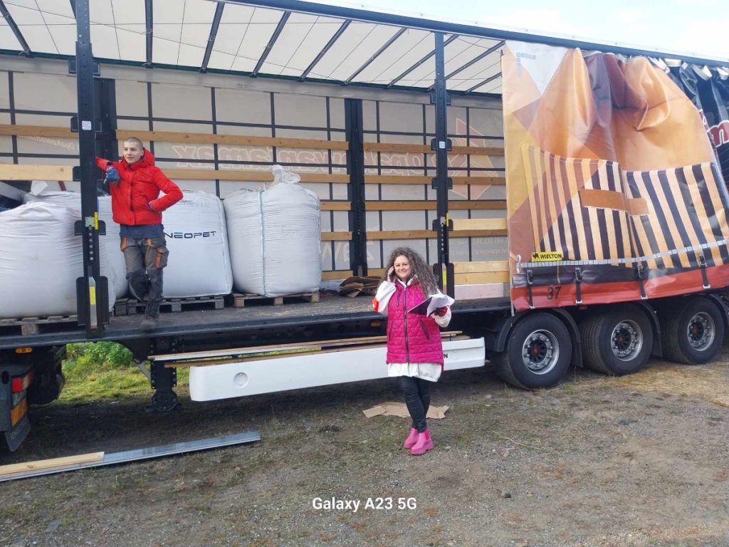 Widok darów dla powodzian na samochodzie ciężarowym. Na środku zdjęcia Pani odbierająca dary, na pace ciężarówki osoba przekazująca.