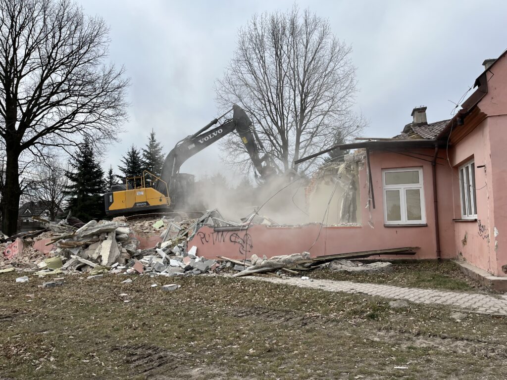 Wyburzanie budynku po szkole w Janiszewie. Widać koparkę, część stojącego budynku oraz gruzy.