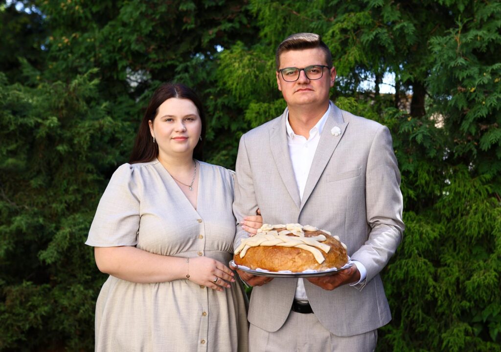 Dwoje osób-od prawej kobieta i obok mężczyzna trzymający w dłoniach chleb.