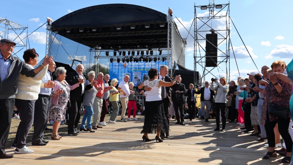 Grupa tańczących osób - mężczyzn i kobiet na przygotowanych dechach. Wszyscy tańczą tworząc koło a po środku para.