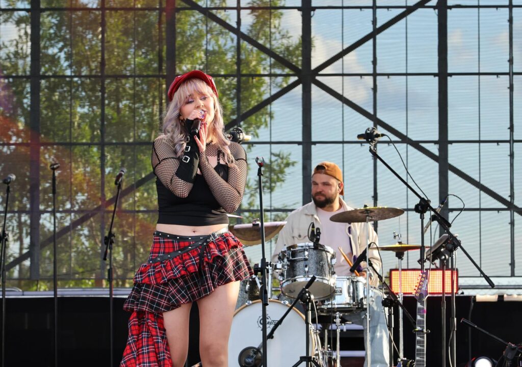 Dziewczyna śpiewa trzymając mikrofon. W tle mężczyzna gra na perkusji.