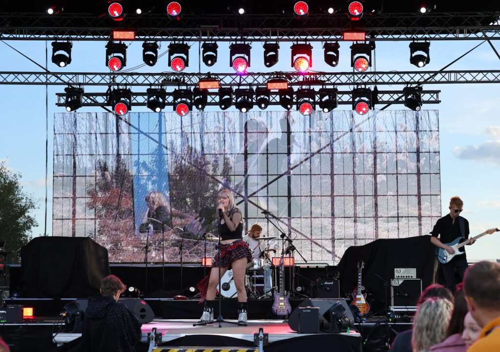 Dziewczyna śpiewa trzymając mikrofon. W tle mężczyźni grają na instrumentach. Widać scenę z oświetlaniem.