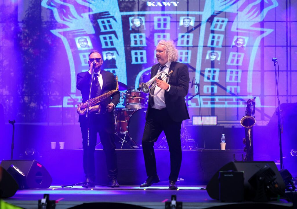 Na scenie dwaj mężczyźni grający na instrumentach muzycznych. W tle kolorowa grafika.