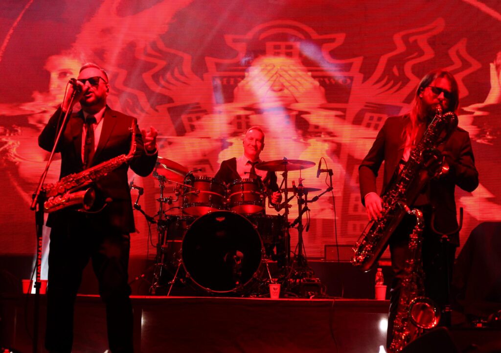 Na scenie trzej mężczyźni grający na instrumentach muzycznych a pierwszy od prawej śpiewa do mikrofonu. W tle kolorowa grafika.