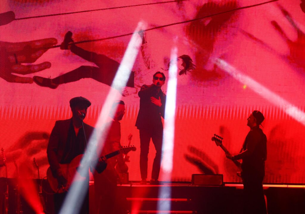 Po środku sceny śpiewa do mikrofonu mężczyzna. Po bokach dwaj mężczyźni grający na instrumentach. W tle kolorowa grafika.