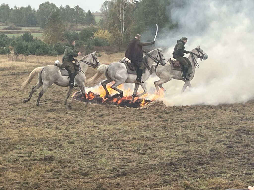 Inscenizacja pokazów kawaleryjskich - trzej jeźdźcy na koniach, konie przeskakują przez ognisko, nad którym unosi się dym. W tle drzewa.