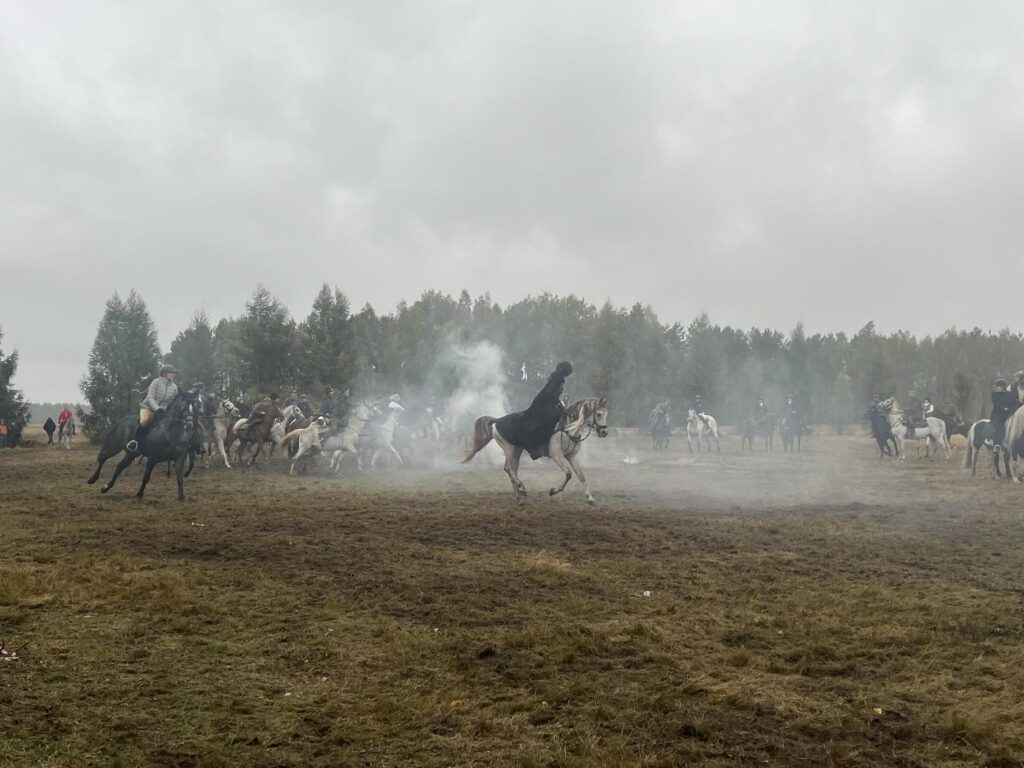 Duża grupa jeźdźców na koniach  podczas XIII Hubertusa. W tle drzewa i dym z ogniska.