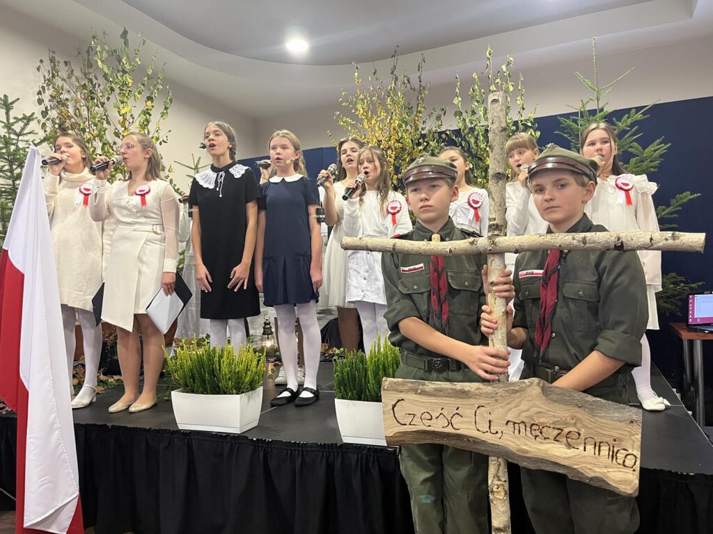 Grupa młodzieży w inscenizacji przedstawienia o Teresie Grodzińskiej. Po lewo flaga Polski. Po prawo dwaj harcerze z drewnianym krzyżem.