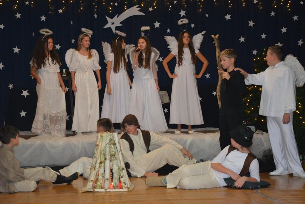 Na zdjęciu przedstawiona jest inscenizacja jasełek, czyli przedstawienia bożonarodzeniowego, przygotowanego przez dzieci i młodzież. Scena udekorowana jest w świątecznym klimacie: w tle widać granatową kurtynę z gwiazdami, świecące lampki oraz choinki, co tworzy nocną, betlejemską scenerię.

W centrum stoją aniołowie ubrani w białe szaty, z aureolami i skrzydłami. Przed nimi leża lub siedzą pasterze oraz po prawo stoją diabeł i anioł. Dzieci odgrywające role mają starannie dobrane kostiumy, co podkreśla uroczysty i podniosły charakter przedstawienia.



