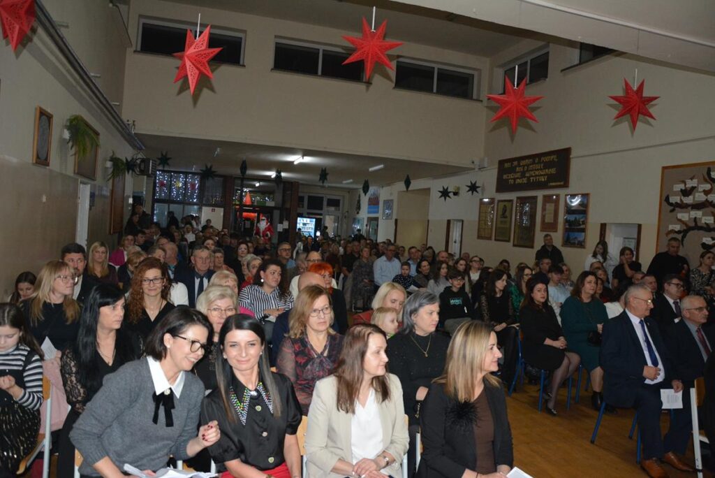 Na zdjęciu widać zgromadzoną publiczność podczas wieczoru wigilijnego odbywającego się w holu PSP w Zakrzewie. W pierwszym rzędzie siedzą dorośli – kobiety ubrane w eleganckie, formalne stroje (żakiety,  sukienki), co sugeruje oficjalny charakter wydarzenia. Wśród nich można zauważyć przedstawicieli lokalnych władz i zaproszonych gości.
Atmosfera zdjęcia jest podniosła i oficjalna.