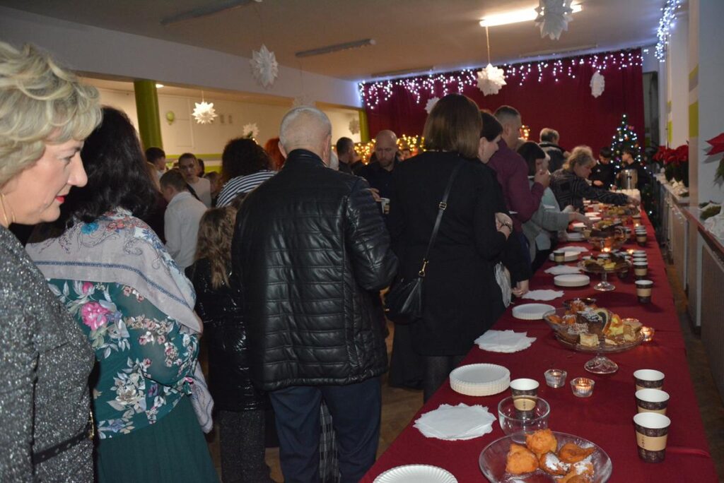 Na zdjęciu od prawej strony widać stoły z wigilijnymi potrawami a po lewo grupa ludzi częstujących się. 