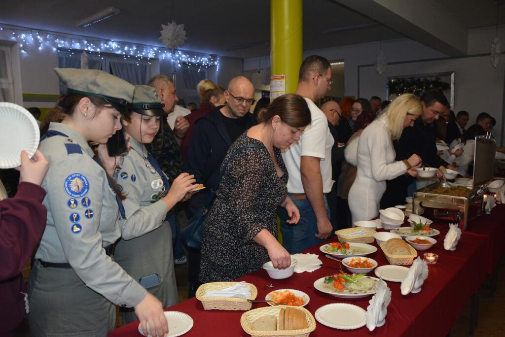 Na zdjęciu od prawej strony widać stoły z wigilijnymi potrawami a po lewo grupa ludzi częstujących się. 