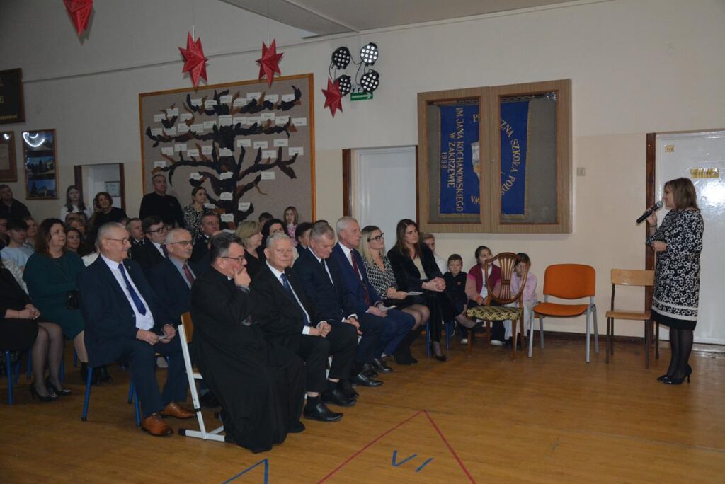 Na zdjęciu widać zgromadzoną publiczność podczas wieczoru wigilijnego odbywającego się w holu PSP w Zakrzewie. W pierwszym rzędzie siedzą dorośli – kobiety i mężczyźni ubrani w eleganckie, formalne stroje (garnitury, marynarki, sukienki), co sugeruje oficjalny charakter wydarzenia. Wśród nich można zauważyć osoby duchowne oraz przedstawicieli lokalnych władz lub zaproszonych gości.

Po prawej stronie kadru stoi kobieta przemawiająca do zgromadzonych, trzymająca mikrofon. Za nią znajdują się krzesła oraz dzieci siedzące przy ścianie, obserwujące wydarzenie. Na ścianach wiszą szkolne tablice, sztandary oraz dekoracje, w tym czerwone gwiazdy i tablica pamiątkowa.

Atmosfera zdjęcia jest podniosła i oficjalna.