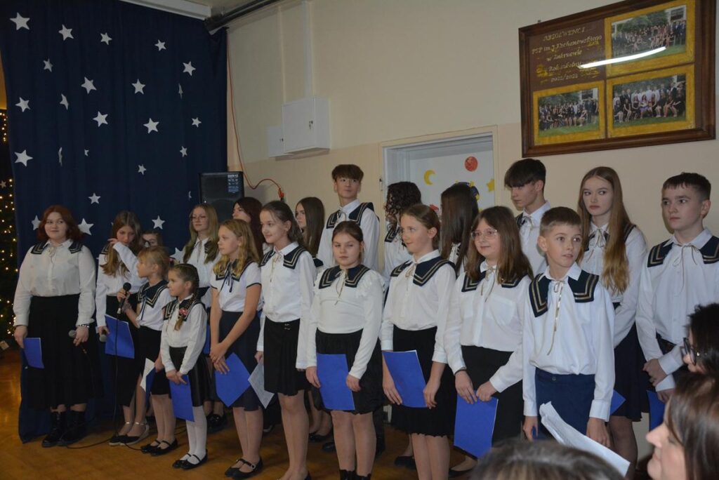 Na zdjęciu widać grupę dzieci i młodzieży stojących w kilku rzędach podczas wieczoru wigilijnego. Uczestnicy są ubrani w jednakowe, galowe stroje: białe koszule z granatowymi kołnierzami w marynistycznym stylu oraz ciemne spódnice lub spodnie. Wielu z nich trzyma w rękach niebieskie kartki z tekstem.

Scena znajduje się w holu PSP w Zakrzewie. Po lewej stronie widać granatową kurtynę z motywem białych gwiazd, a w tle świąteczne lub dekoracyjne oświetlenie. Na ścianie po prawej stronie wiszą oprawione zdjęcia grupowe. Atmosfera zdjęcia jest oficjalna, ale jednocześnie uroczysta i spokojna.