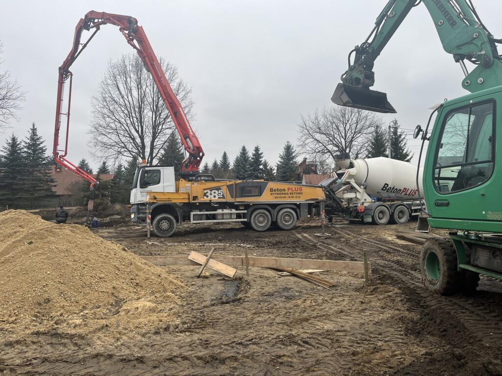 Plac budowy z wykopami fundamentowymi, na którym pracuje duża czerwona pompa do betonu zamontowana na ciężarówce. Kilku pracowników w odzieży roboczej nadzoruje wylewanie betonu. W tle widać drzewa iglaste, trawnik oraz kilka domów jednorodzinnych pod pochmurnym niebem