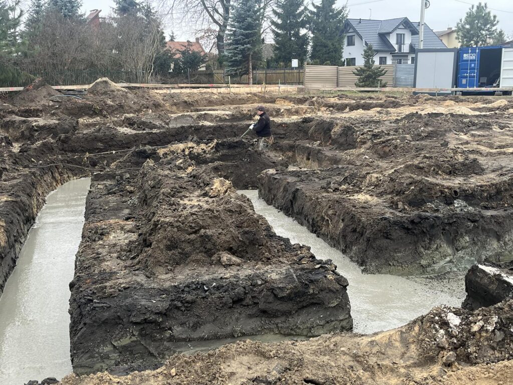 Plac budowy z głębokimi wykopami wypełnionymi świeżo wlanym betonem. Między wykopami stoi pracownik w odzieży roboczej. W tle widać sąsiednie domy, ogrodzenia oraz kontenery budowlane