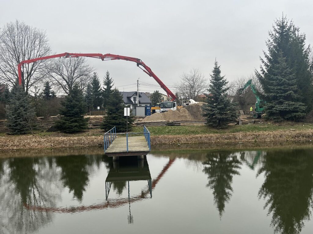Widok przez staw na budowę, z niebieskim pomostem na pierwszym planie. Za nim pracuje czerwona pompa do betonu oraz zielona koparka, a obok leżą stosy materiałów i kopce piasku. W tle rosną świerki i widoczne są pobliskie domy.