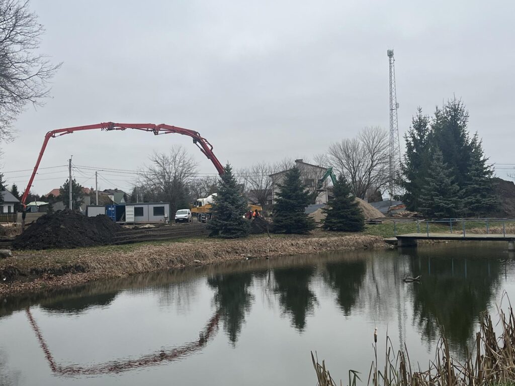 Budowa widoczna po drugiej stronie stawu; duże ramię czerwonej pompy do betonu rozciąga się nad placem. Na terenie stoją kontenery, pojazdy budowlane i kopce ziemi. W tle znajdują się drzewa, domy oraz wysoki maszt, a w tafli wody odbijają się maszyny i krajobraz