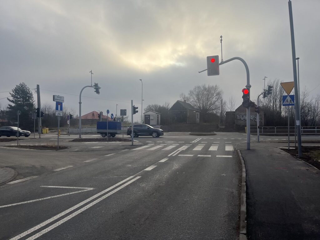 Dzienne zdjęcie tego samego skrzyżowania. Sygnalizacja świetlna wyświetla czerwone światło. Widoczne są samochody w oddali, znaki drogowe, przejście dla pieszych i zabudowania w tle