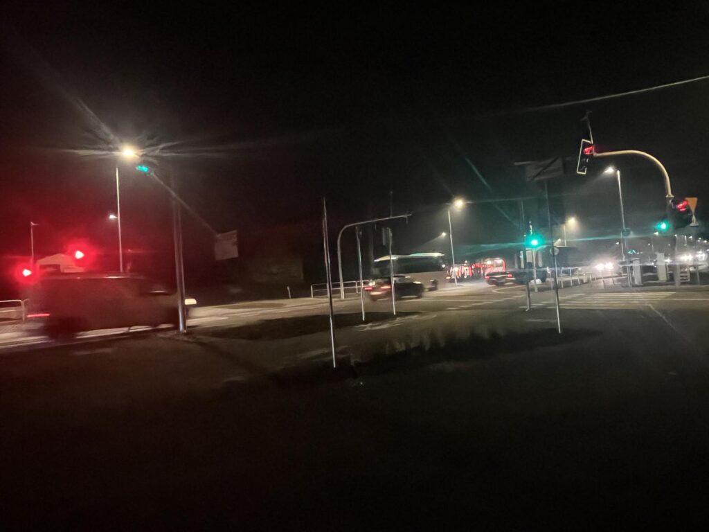 Nocna scena przedstawiająca dobrze oświetlone skrzyżowanie z sygnalizacją świetlną. Na pierwszym planie widać rozmyty samochód przejeżdżający obok czerwonych świateł. W tle znajdują się inne pojazdy, w tym autobus, poruszające się w wielu kierunkach przy zielonych światłach. Latarnie uliczne oświetlają okolicę.