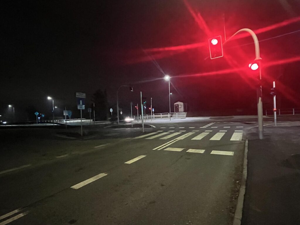 Nocne zdjęcie pustego skrzyżowania. Sygnalizacja świetlna pokazuje czerwone światło w kierunku fotografa. Widać pasy jezdni, przejście dla pieszych oraz latarnie oświetlające otoczenie
