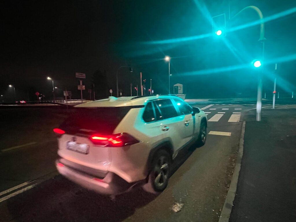 Nocna scena skrzyżowania z sygnalizacją świetlną. Jasny SUV jedzie przez skrzyżowanie przy zielonym świetle. W tle widać latarnie uliczne, znaki drogowe i pasy przejścia dla pieszych