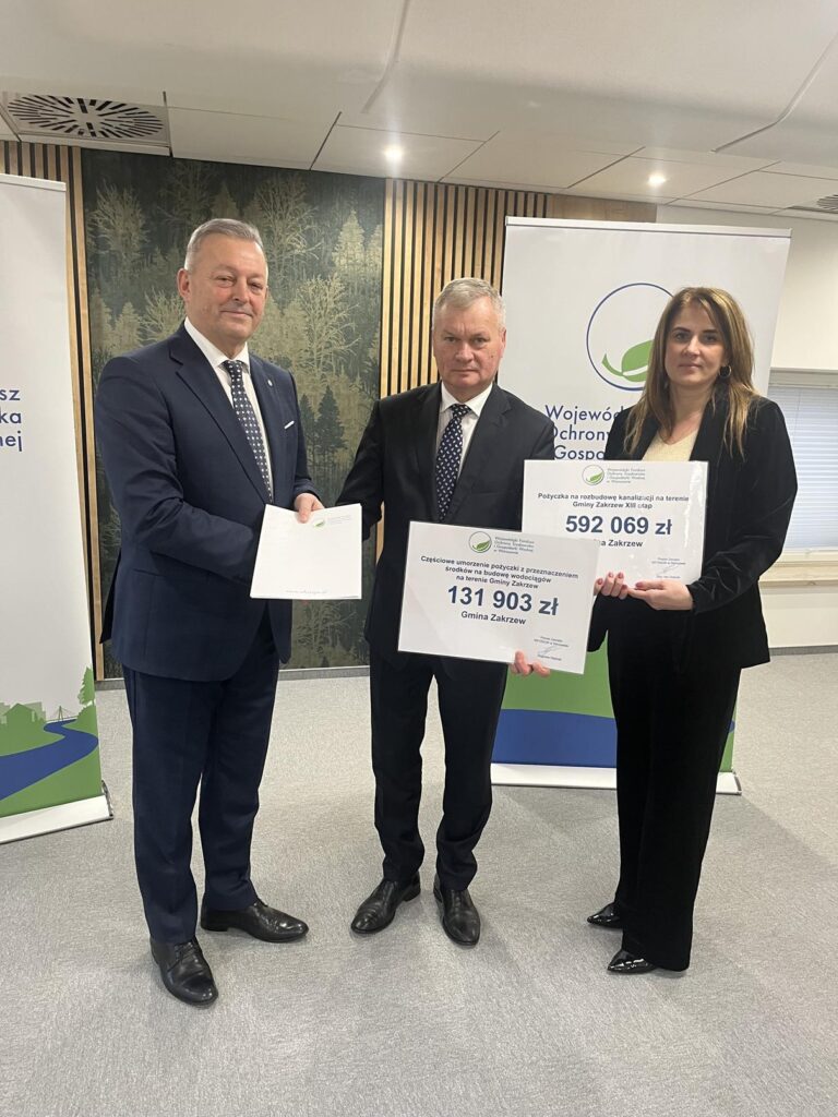 Trzy osoby w eleganckich strojach stoją w pomieszczeniu konferencyjnym. Prezes Zarządu WFOŚiGW wręcza dokument Wójtowi Gminy Zakrzew, który trzyma czek o wartości 592 069 zł na częściowe umorzenie pożyczki, Skarbnik Gminy Zakrzew po prawej trzyma czek o wartości 131 903 zł na budowę kanalizacji w gminie Zakrzew. W tle stoją roll-upy Wojewódzkiego Funduszu Ochrony Środowiska i Gospodarki Wodnej w Warszawie.