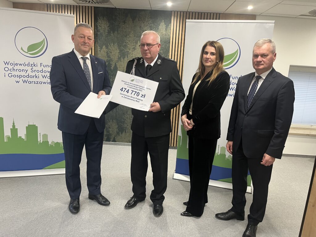 Z lewej strony Prezes Zarządu WFOŚiGW trzyma dokumenty, strażak w galowym mundurze – trzyma duży symboliczny czek na kwotę 474 770 zł dla Ochotniczej Straży Pożarnej w Zakrzewskiej Woli. Obok nich stoi Wójt Gminy Zakrzew oraz P. Skarbnik. W tle znajdują się roll-upy Wojewódzkiego Funduszu Ochrony Środowiska i Gospodarki Wodnej w Warszawie.