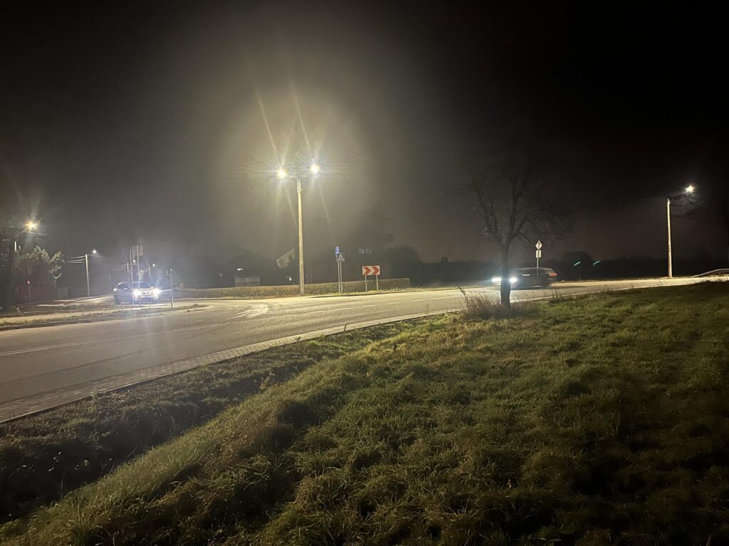 Na zdjęciu widać ciche skrzyżowanie nocą, oświetlone wysokimi latarniami ulicznymi. Powietrze wydaje się mgliste lub zamglone, co tworzy aureole i efekty rozbłysków wokół świateł. Widocznych jest kilka samochodów z włączonymi światłami – jeden nadjeżdża z lewej, a drugi z prawej – a na pierwszym planie widać trawiasty nasyp przydrożny. Znajdują się tam znaki drogowe wskazujące na zakręt lub rondo, a sylwetki drzew i budynków znikają w ciemnym tle.