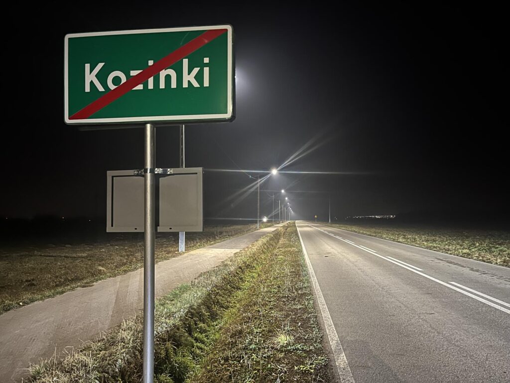 Na zdjęciu widać na pierwszym planie zielony znak miejski z napisem „Koźinki” przekreślony ukośnym czerwonym pasem, który zazwyczaj oznacza wyjazd z miejscowości. Znak jest zamontowany na metalowym słupie, a pod nim znajduje się kolejny, mniejszy znak.
Po prawej stronie rozciąga się prosta, dwupasmowa droga oświetlona równomiernie rozmieszczonymi latarniami, które tworzą jasne plamy światła i długie odbicia na asfalcie. Po lewej stronie znajduje się wąski chodnik lub ścieżka rowerowa, biegnąca równolegle do drogi, oddzielona pasem trawy. Okolica wygląda na wiejską lub półwiejską – otwarte pola, niewiele budynków i odległe światła na horyzoncie. Niebo jest ciemne i lekko zamglone, co podkreśla blask latarni.