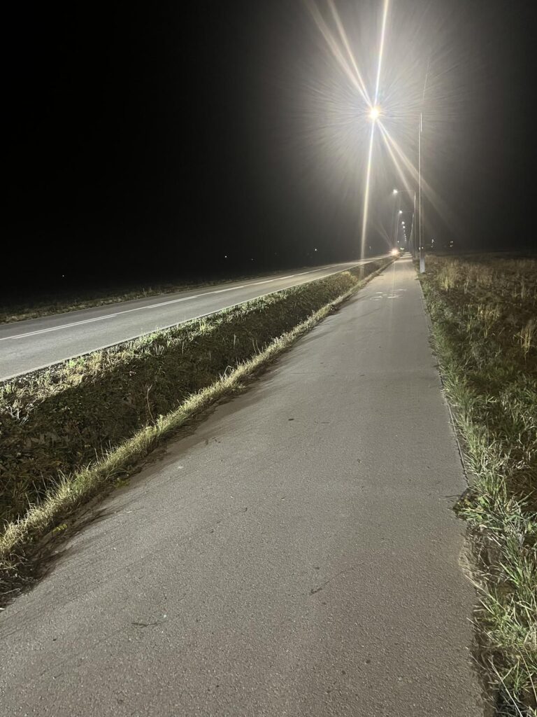 Na zdjęciu widać cichą, pustą drogę nocą. Prosta, utwardzona ścieżka (prawdopodobnie chodnik lub ścieżka rowerowa) biegnie wzdłuż drogi.
Wysokie latarnie uliczne oświetlają ścieżkę, tworząc silne rozbłyski światła w ciemnym, mglistym powietrzu.
Okolica wygląda na wiejską lub podmiejską, z trawą i otwartymi polami po obu stronach.
Nie widać żadnych ludzi, a jedynie światło odległego pojazdu w oddali, co dodaje spokojnej, nieco upiornej atmosfery.