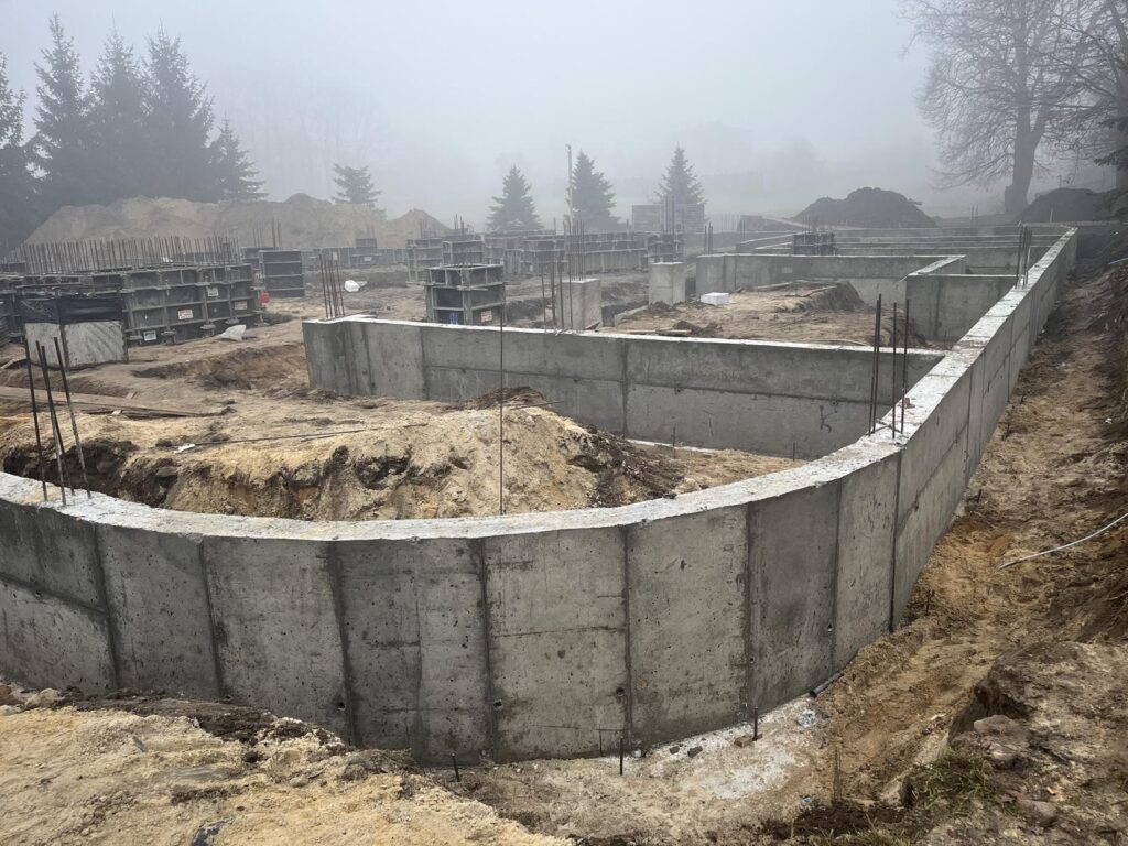 Plac budowy z wylanymi betonowymi ścianami fundamentowymi i wystającym zbrojeniem; w tle mgła, drzewa i hałdy ziemi.