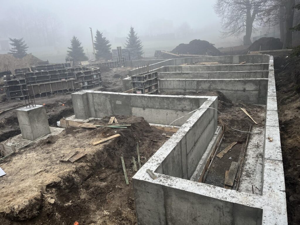 Widok placu budowy z betonowymi ścianami fundamentowymi tworzącymi zarys budynku; w wykopach ziemia i deski, w tle szalunki, hałdy gruntu oraz drzewa we mgle.