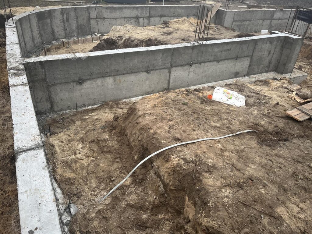 Zbliżenie na betonowe ściany fundamentowe na placu budowy; przed nimi wykop z ziemią i przewodem, wewnątrz fundamentów nasyp gruntu i wystające pręty zbrojeniowe.