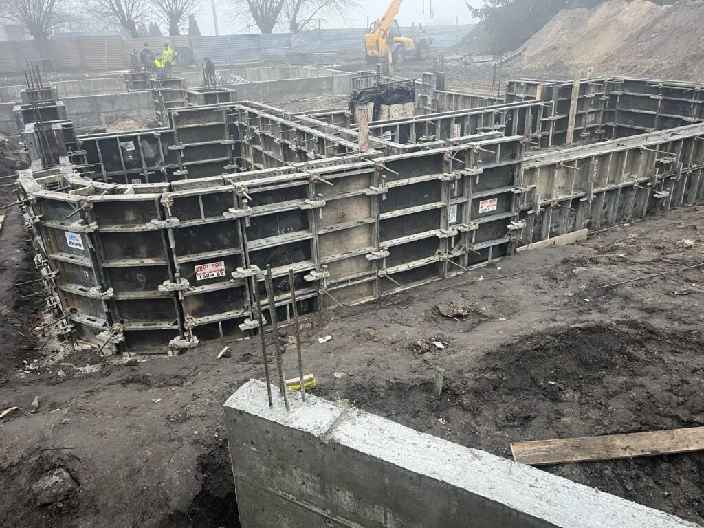 Plac budowy z widocznym szalunkiem stalowym pod fundamenty budynku. W wykopie stoją zamontowane formy do betonowania ścian i ław. Całość znajduje się na etapie przygotowania fundamentów.