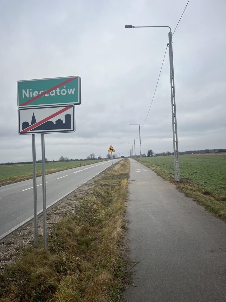 Na zdjęciu widać na pierwszym planie zielony znak miejski z napisem „Nieczatów” przekreślony ukośnym czerwonym pasem, który zazwyczaj oznacza wyjazd z miejscowości. Znak jest zamontowany na metalowym słupie, a pod nim znajduje się kolejny, mniejszy znak.
Po prawej stronie rozciąga się prosta, dwupasmowa droga z równomiernie rozmieszczonymi latarniami. Po lewej stronie znajduje się wąski chodnik lub ścieżka rowerowa, biegnąca równolegle do drogi, oddzielona pasem trawy. Okolica wygląda na wiejską lub półwiejską – otwarte pola, niewiele budynków.