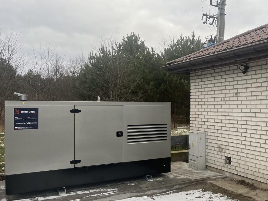 Agregat prądotwórczy w metalowej obudowie z logo Enerwigo, zainstalowany na zewnątrz przy budynku technicznym, w otoczeniu drzew i infrastruktury energetycznej