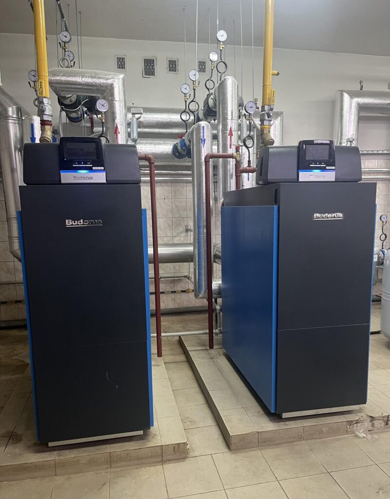 Dwa stojące kotły grzewcze Buderus ustawione obok siebie w kotłowni, podłączone do instalacji rurowej i gazowe