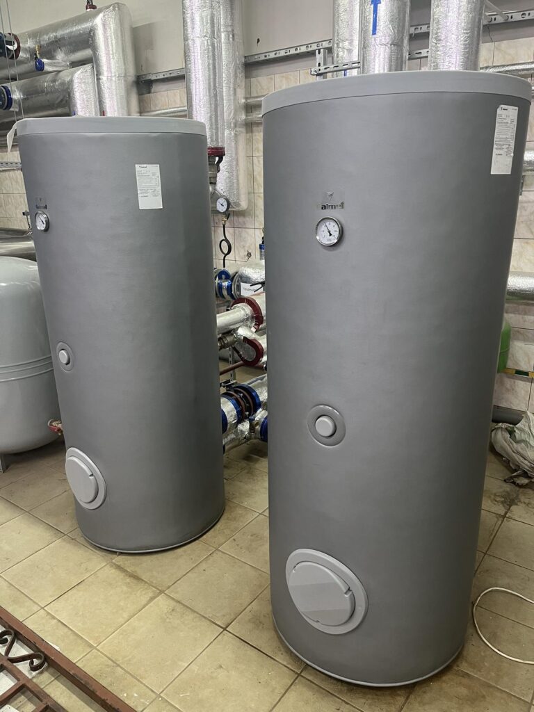 Dwa pionowe, cylindryczne zbiorniki akumulacyjne ciepłej wody z wbudowanymi termometrami, stojące w pomieszczeniu technicznym