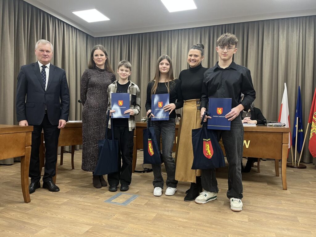 Sesja Rady Gminy w Zakrzewie-wręczenie nagród młodym sportowcom.
Na zdjęciu widać salę konferencyjną z drewnianymi podłogami, zasłonami zasłaniającymi tylną ścianę i jasnymi lampami sufitowymi. Za grupą znajduje się długi, drewniany stół z mikrofonami i dokumentami.
Po lewej stronie stoi Wójt Gminy Zakrzew w ciemnym garniturze i krawacie. Obok niego stoją trzy młode osoby, ubrane w stroje codzienne lub półformalne (ciemne topy, dżinsy, trampki, trzymają granatowe urzędowe teczki z herbem gminy i torby upominkowe ), pomiędzy dziećmi stoją dwie dorosłe kobiety.
Za stołem, po prawej stronie, siedzi dorosła kobieta, obserwując przebieg wydarzenia.
Po prawej stronie widocznych jest kilka flag, w tym coś, co wygląda na flagę narodową, flagę Unii Europejskiej oraz flagę regionalną.